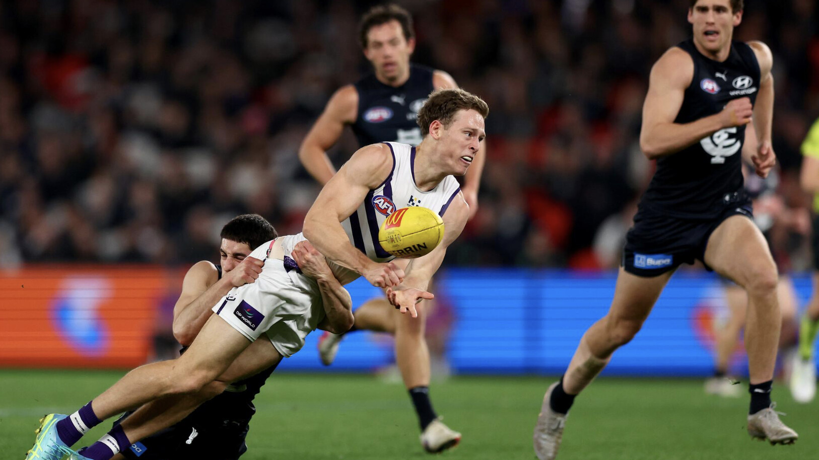 A Fremantle player fires out a handball as a Carlton tackler wraps him up at Marvel Stadium